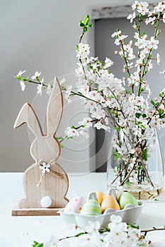 Wooden Easter bunny with blooming tree branch with flowers in the vase and colored easter eggs in container on the white