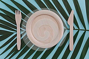 Wooden empty plate, fork and knife on palm leaf background on blue. Zero waste concept