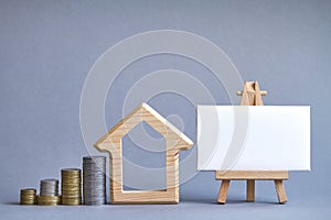 Wooden figure of house with several columns of coins nearby and white board on miniature easel on gray background, the concept of