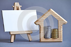 Wooden figure of house with two columns of coins inside and white board on miniature easel on gray background, the concept of
