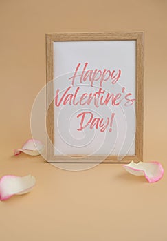 Wooden frame with text HAPPY VALENTINES DAY on paper blank and delicate pink rose petals on beige background. Romantic