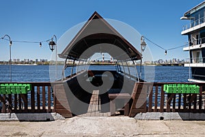 A wooden gazebo in the shape of a boat with a deck and a sail roof with lanterns near the river