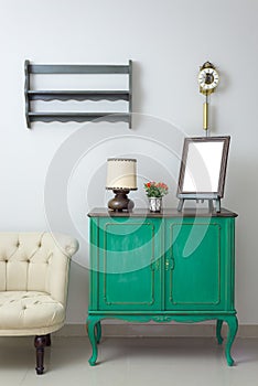 Wooden green vintage sideboard with empty wooden ornate brown desktop photo frame, flower planter, table lamp and cream tub chair