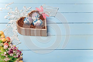 Wooden heart shaped box with decorated Easter eggs. White lace, flowers, pink dragonfly toy on blue background.
