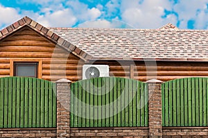 Wooden house with air conditioning, surrounded by a green fence. Blue sky with clouds. General plan