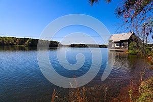 A wooden house on the banks of the lake with vast blue lake water surrounded by lush green trees and plants with powerful clouds