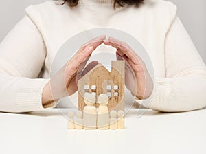 A wooden house and figures of a family, two female palms protect objects from above. Real estate insurance concept, happy family