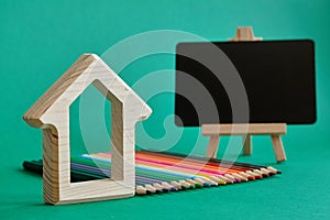 Wooden house figurine, miniature chalk board on an easel and color pencils laid out by rainbow colors isolate on green background