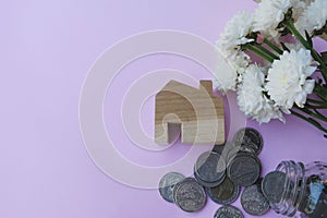 Wooden house model, coins flowing out from glass jar on pastel pink background. House finance, home mortgage, property investment