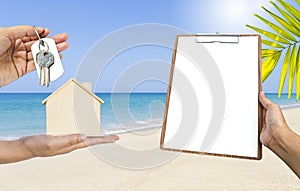 Wooden house model with key in girl hand and blank clipboard over tropical beach