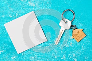 Wooden house shaped keychain and key with white empty paper on blue wooden background