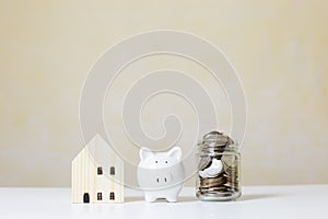 Wooden house with white piggy bank and money coins in jar glass on table, property investment,  saving for future.