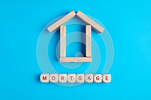 Wooden house and the word mortgage formed with wooden blocks on blue background. Conept of purchasing a house with mortgage