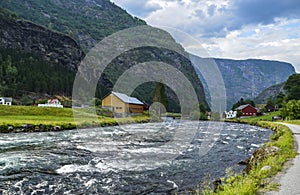 Wooden houses and clear river at the foot of a mountain covered with green grass. Summer scene in Norway