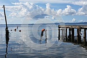 Wooden jetty for boats against a blue sky with some clouds. Floating buoys and fishing nets. Hills on background. Bolsena lake,