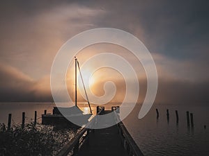 Wooden jetty with a sailboat in the water of a lake in fog at sunrise