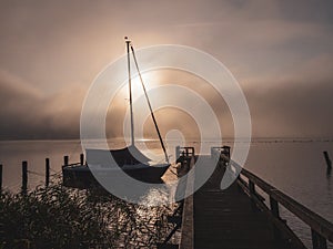 Wooden jetty with a sailboat in the water of a lake in fog at sunrise