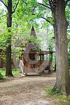 A wooden log hut with a patterned porch among green trees in the forest. An old log house among the trees. A porch with