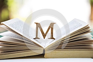 Wooden M font design on open old book over blurred background