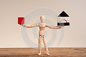 Wooden man comparing two things , hold red cube and black and white mini house.