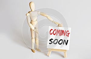 Wooden man shows with a hand to white easel with text COMING SOON