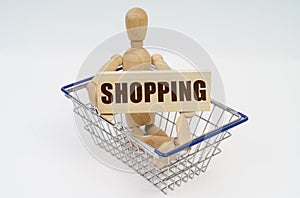 A wooden man sits in a shopping basket, holding a sign in his hands - SHOPPING
