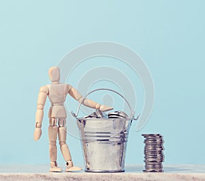 A wooden mannequin and coins in a miniature bucket on a blue background, a concept of high income, subsidy