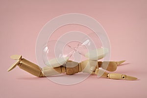 Wooden mannequin lying under hourglass on pink background - Concept of stopping time, health and aging