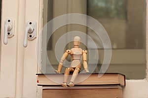 A wooden mannequin sits on a stack of books against the background of an old window.