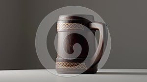 Wooden oak mug with a handle is spinning on white table. Brown mug with ornament isolated on dark gray studio background
