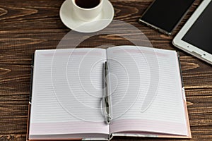 A wooden office desk with many things on it. Diary with pen, Samsung phone. White cup of coffee. Top view with copy space