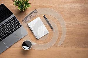 Wooden office desk table with blank notebook, laptop computer, cup of black coffee and supplies. Top view with copy space, flat