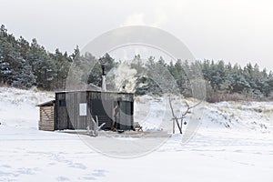 Wooden outdoor sauna with a smoke coming out of chimney on a beautiful cold snowy winter day at the Baltic sea.