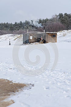 Wooden outdoor sauna with a smoke coming out of chimney on a beautiful cold snowy winter day at the Baltic sea.