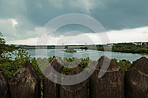 Wooden palisade stands against the backdrop of a hydroelectric dam under a dark stormy sky