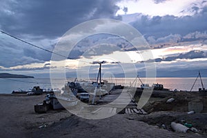 Wooden path to pier with old ships and boats in evening at sunset with blue clouds on lake baikal, mountains on horizon