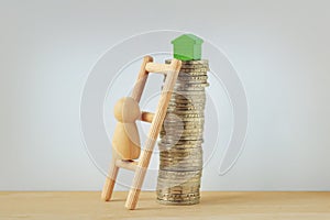 Wooden pawn climbing a ladder to reach a little green house on top of coin tower - Concept of buying home