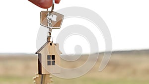 Wooden pendant of a house and key in hand. Background of the sky and field. Dream of home, building a cottage in the countryside,