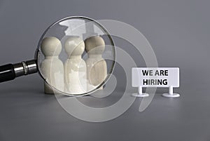 Wooden people with magnifying glass and signboard written with We Are Hiring