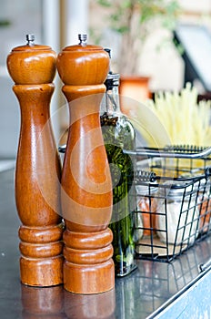 Wooden pepper mills, a bottle of extra virgin olive oil with spices and herbs, salt shakers on on a stainless surface