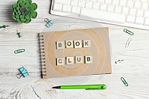 Wooden phrase Book club  in workspace on paper notepad with computer keyboard, supplies  and green plant on light wood background