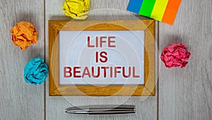 Wooden picture frame with inscription `life is beautiful` on beautiful white wooden table, colored paper and metalic pen.