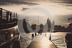 Wooden pier with boats at sunset in Amsterdam dock. Amsterdam city silhouettes in evening dusk light.