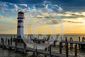 A wooden pier leading to the lake and a red and white lighthouse on Lake Neusiedl in Podersdorf, Austria. In the background is a