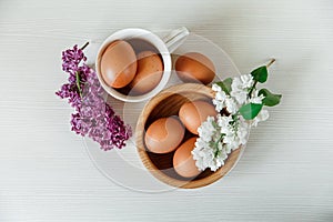 Wooden Plate and Ceramic Cup with Eggs,White and Purple Branches of Lilac.White Wooden Background.Top View