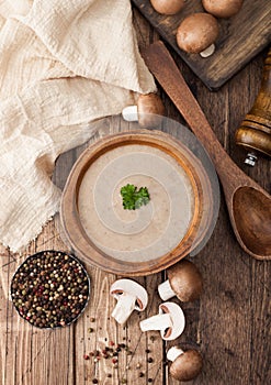 Wooden plate of creamy chestnut champignon mushroom soup with wooden spoon, pepper and kitchen cloth on wooden background. Top