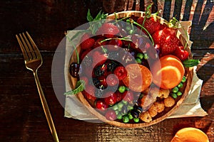 Wooden plate with seasonal fruits and berries background under hard shadows. Strawberries, apricots, cherry, raspberry