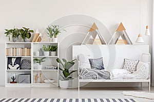 Wooden rack with books, fresh plants and cushions in white bedro
