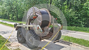 A wooden reel of insulated electrical cable is mounted on a metal two-axle trailer with leaf springs, which stands on a tile sidew