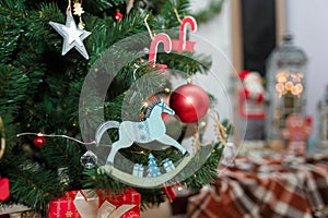 wooden rocking horse toy and decorations hanging on fir tree branches with santa claus and lantern lights on blurry background.